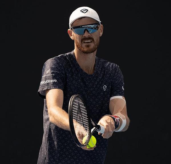 Male tennis player in dark shirt and white cap preparing to serve with racket and ball.